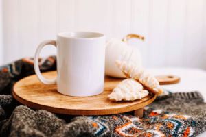 A cup of coffee, a white ceramic pumpkin, and a couple of treats sitting on a wooden cutting board atop a warm woolen sweater.